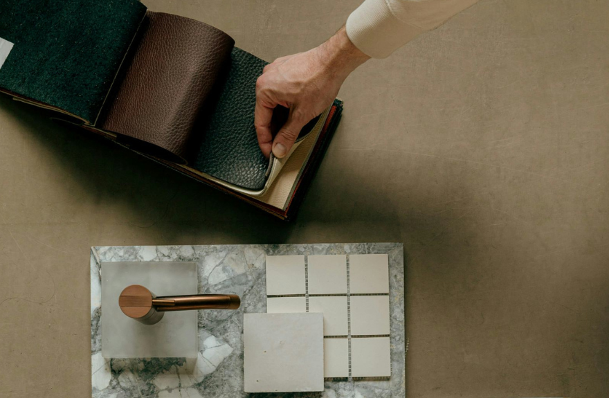 A hand flipping through a leather swatch book, showing different textures and colors, placed next to a marble surface with several tiles and a metal object, likely used for design or material selection